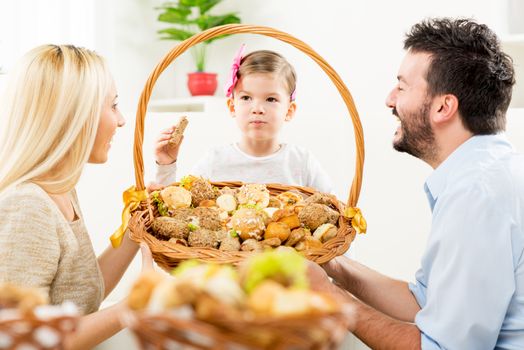 Young parents kneel in front of their little daughter with a smile watching her eat pastries from a large woven basket which together hold in their hands.