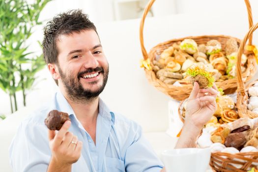A young man with a beard, holding in his hands a sweet and savory pastries. With a smile looking at the camera. In the background you can see woven baskets with pastries.