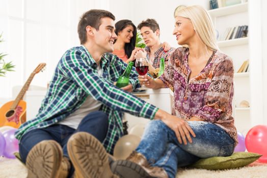 Young couple at a home party, sit on the floor, knocking the glasses toasting, in the background you can see another couple.