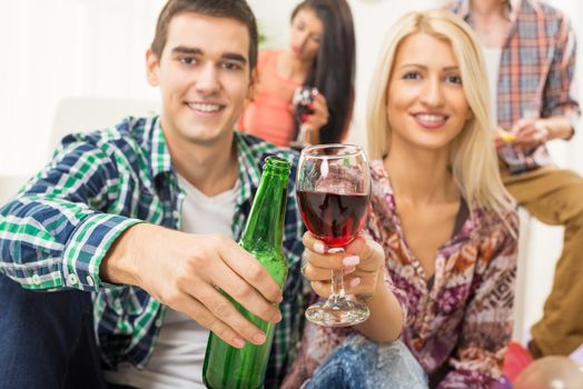 Young couple at home party with a smile on their faces out of focus, toasting with drinks that are in the foreground. In the background is another young couple.