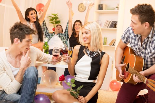 A young guy is courting the pretty girl at home party, while their friend plays acoustic guitar, and in the background the rest of society with arms raised celebrated their relationship.