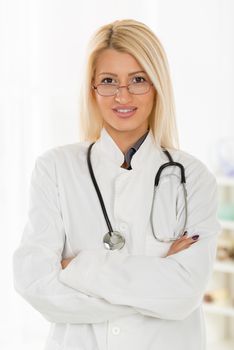 Young cute blonde female  doctor with the glasses, dressed in a white coat standing arms crossed with a stethoscope over the neck, with a smile looking at the camera.