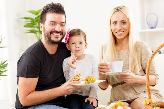 Happy family in front of the wicker basket filled with tasty baked goods. Cute little girl sitting in daddys lap, next to her mother, holding a bun, a mother holding a cup.