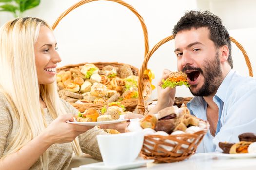 Beautiful blonde girl feeds her man with a pastry. In the background you can see woven baskets with bakery products.