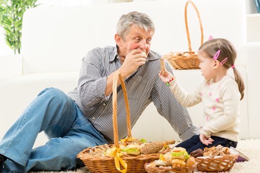Grandfather and granddaughter sitting on a carpet in the living room next to woven baskets with pastries. They look at each other and eat pastry.