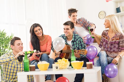 A small group of young people at the home party, enjoy the sounds of the acoustic guitar, drinking wine and beer.