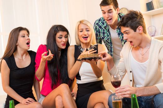 A small group of young people at a birthday party, sitting on the couch, with cheerful expressions on the faces looking at birthday cake in the hands of an attractive blond girl.