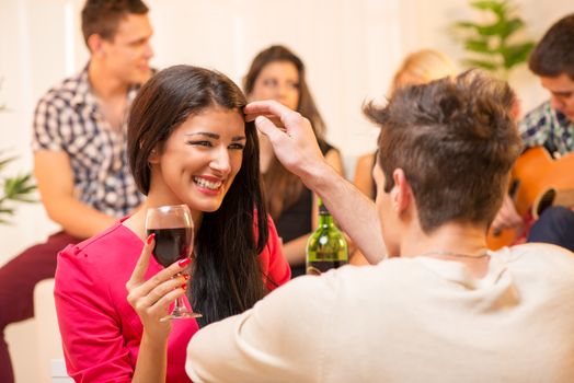The young man at the house party is courting the pretty brunette caressing her hair, and in the background you can see a small group of young people having fun at a party.
