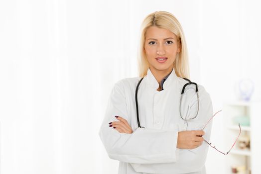 Beautiful young blonde woman in a white coat with a stethoscope over the neck, standing arms crossed, holding glasses and smiling looking at the camera.