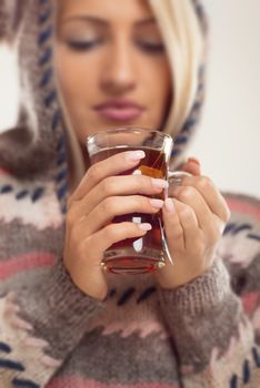 Close-up cup of tea held by the hands of girl, with a face out of focus, dressed in wool sweater with hood.