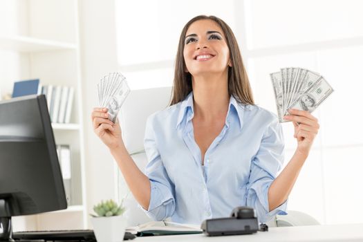 Young businesswoman in office, sitting at an office desk, holding money and with an expression of happiness on her face looking up.