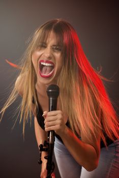 A young woman rock singer with tousled long hair holding a microphone with stand and sing with a wide open mouth.