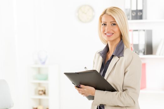 Young blonde businesswoman standing in office, holding folder, smiling looking at the camera. In the background you can see the shelves with binders.