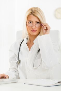 Attractive blonde female doctor in her consulting room, sitting at the table in front of laptop and looking at the camera, underneath her glasses.