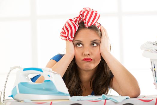Pensive housewife daydreaming leaning on an ironing board holding her head. In front of her are iron and laundry basket.