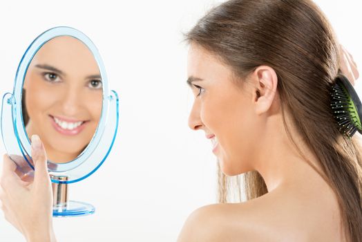 Girl holding a mirror in which looks and combing her long brown hair. Reflection of girl in the mirror looking at the camera with a smile.