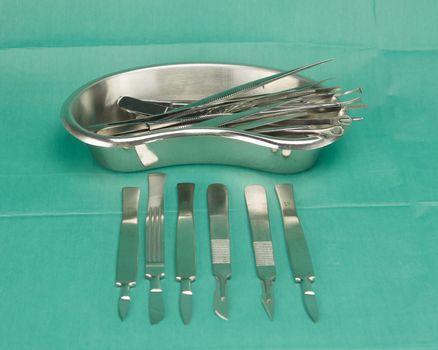 Forceps in kidney shape bowl and scalpel on green fabric.