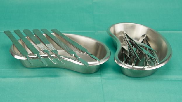 Many types of forceps and scalpel in kidney shape bowl on green fabric.