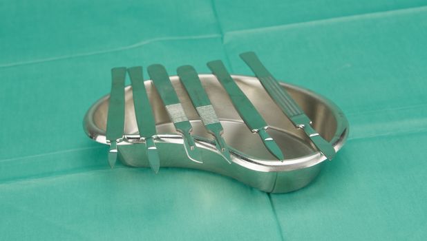 Many types of scalpel in kidney shape bowl on green fabric.