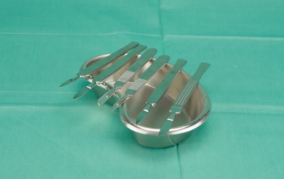 Many types of scalpel in kidney shape bowl on green fabric.