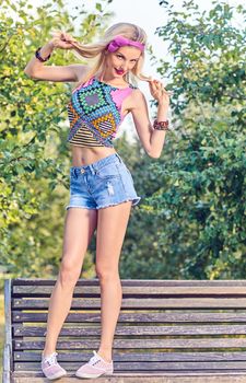 Beauty stylish playful woman on bench, park, people, outdoors.Attractive hipster happy pretty blonde girl with bow, fashionable top, denim shorts. Relax in summer garden,lifestyle