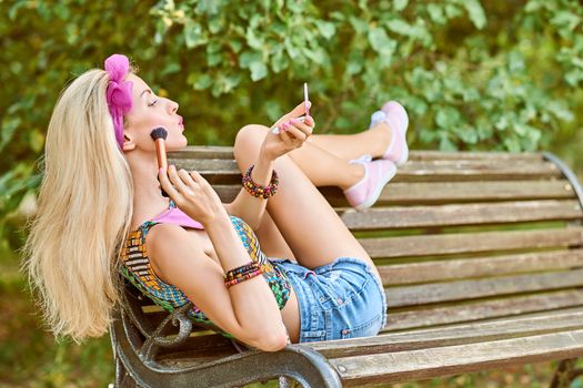 Beauty stylish playful woman primping with mirror on bench, park, people, outdoors.Attractive hipster happy pretty blonde girl with bow, fashionable top, denim shorts. Relax in summer garden,lifestyle