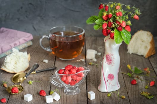 Strawberries in a glass Cup and a bouquet of wild strawberries in a vase. White bread crumb, antique spoons and tea, rustic Style, background blur.