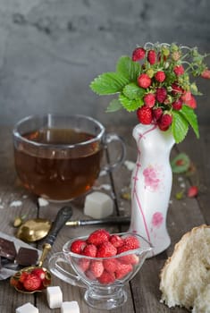 Strawberries in a glass Cup and a bouquet of wild strawberries in a vase. White bread crumb, antique spoons and tea, rustic Style, background blur.