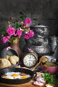 Bacon and eggs in an old pan on the Board. Chopped bacon and cucumbers in a bowl. In the background, old iron pots, broken clock, and a bouquet of tea roses in an old jug. Lies the pot with boiled potatoes, a Cup of milk and bread
