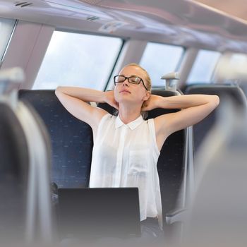 Businesswoman traveling by train, streching on her seat while working on laptop. Business travel concept.