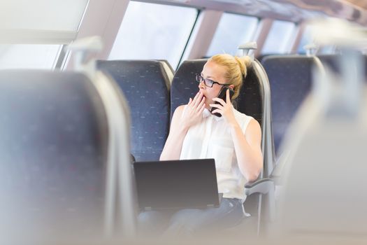 Businesswoman traveling by train, yawning while talking on cellphone and working on laptop. Long and tiresome business travel concept.
