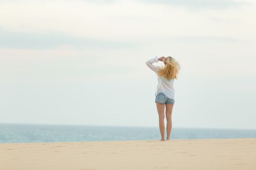 Meditative, sensual blonde woman wearing white loose casual shirt on vacations looking at horizon at dusk.