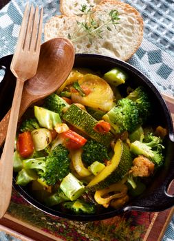 Tasty Colorful Vegetables Ragout with Zucchini, Carrots, Broccoli, Leek and Red Bell Pepper in Black Iron Stewpot with Wooden Spoon and Fork and Bread with Thyme closeup on Blue Napkin