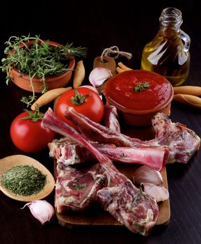 Perfect Raw Lamb Ribs with Spices, Vegetables, Tomato Sauce, Herbs and Small Bottle of Olive Oil on Wooden Cutting Board closeup on Dark Wooden background