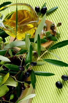 Arrangement of Raw Green and Black Olives with Leafs and Olive Oil in Glass Gravy on Wooden Plate closeup on Green Textile background