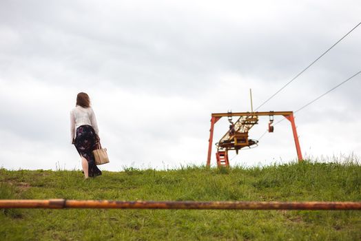 Girl with a satchel in the blouse and skirt rises on grassy Mount Hill. Back vew. Ski elevator as background.