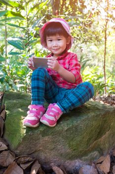Happy asian lovely girl using digital smart phone on boulder, child looking at camera over nature background. Outdoors in the day time with bright sunlight, travel on vacation.