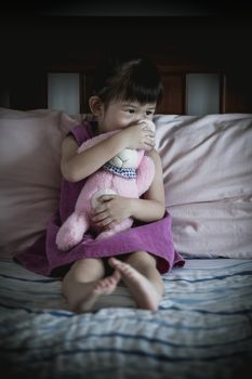 Little sad asian girl sitting on bed with doll in dark bedroom. Vignette picture style.
