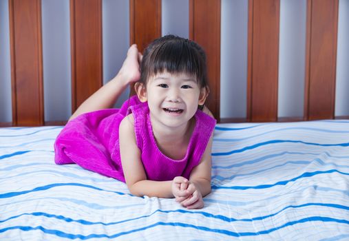 Adorable little asian girl in a purple dress smiling and looking at camera. Cute child  lying barefoot on bed in bedroom.