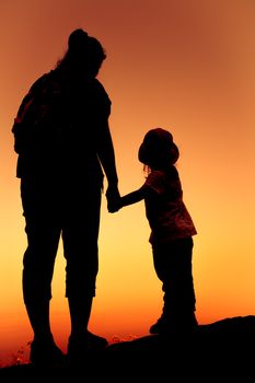 Silhouette back view of mother and daughter clasping hand together at sunset on vacation. Colorful sky background orange and gold colors sunset sky. Friendly family.