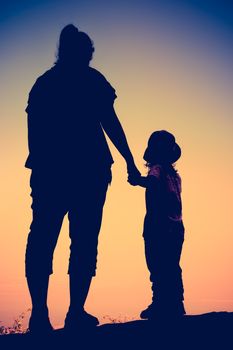 Silhouette back view of mother and child hikers enjoying the view at the top of a mountain. Colorful sunset sky background. Friendly family. Cross process. Vintage style.