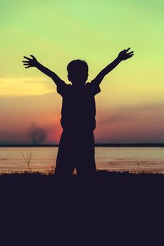 Silhouette back view of child enjoying the view at riverside. Colorful sunset sky background. Boy raising his hands up. Cross process. Vintage style.