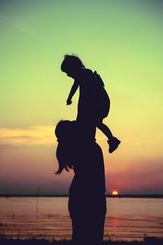 Silhouette of mother and child enjoying the view at riverside. Mother lifting her little girl up in the air on colorful sunset sky background. Friendly family. Cross process. Vintage style.