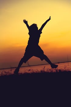 Silhouette back view of child jumping against sunset. Boy enjoying the view at riverside. Happy time on vacation. Vignette style.