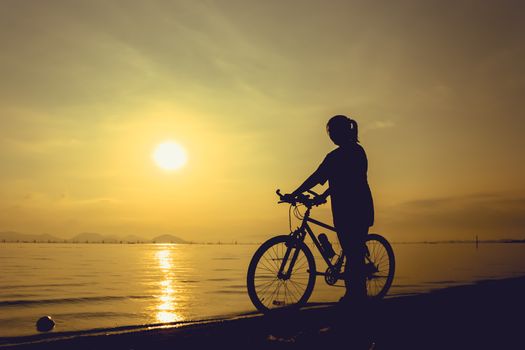 Silhouette of healthy biker-girl enjoying the view at seaside, on colorful sunset yellow sky background. Reflection of sun in water. Active outdoor lifestyle concept. Outdoors.