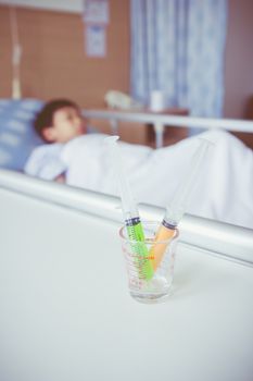 Syringes in a glass measuring cup with blurred illness boy lying on sickbed in hospital. Syringe in focus, child out of focus. Health care and medical concept. Vintage style.
