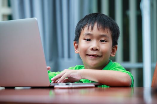 Happy asian child looking at camera. Handsome boy enjoying modern generation technologies playing indoors using laptop computer and mobile phone. Education and learning concept.