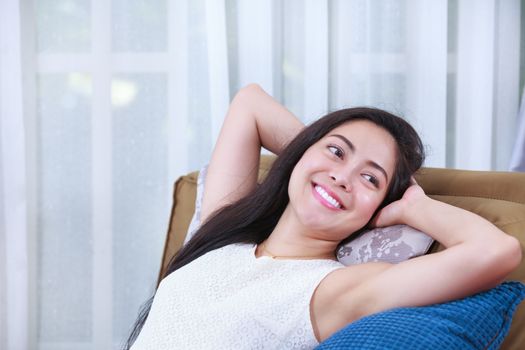 Beautiful asian woman relaxing lying on a couch at home and smiling happy. Resting young female clasping her hands behind her head.