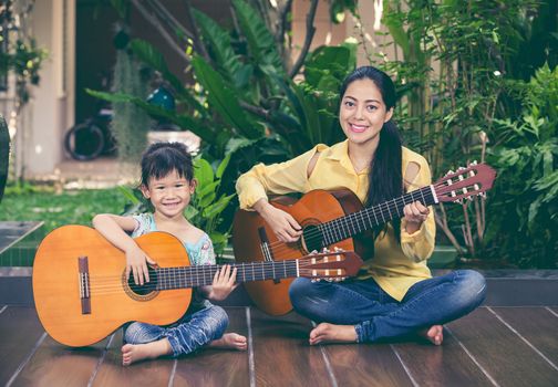 Happy family spending time together at home. Asian mother with daughter playing classic guitar and looking at camera. Vintage picture style. Positive human emotion.