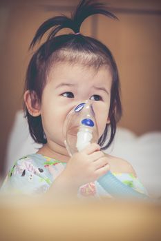 Cute asian child holds a mask vapor inhaler for treatment of asthma on sickbed in hospital. Breathing through a steam nebulizer. Concept of inhalation therapy apparatus. Vintage tone.